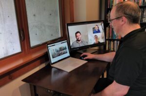 Crewdson participates in a Microsoft Teams meeting from home with a bookshelf in the background, meeting participants displayed on an external monitor, and Microsoft SharePoint displayed on a Microsoft Surface Book 2.