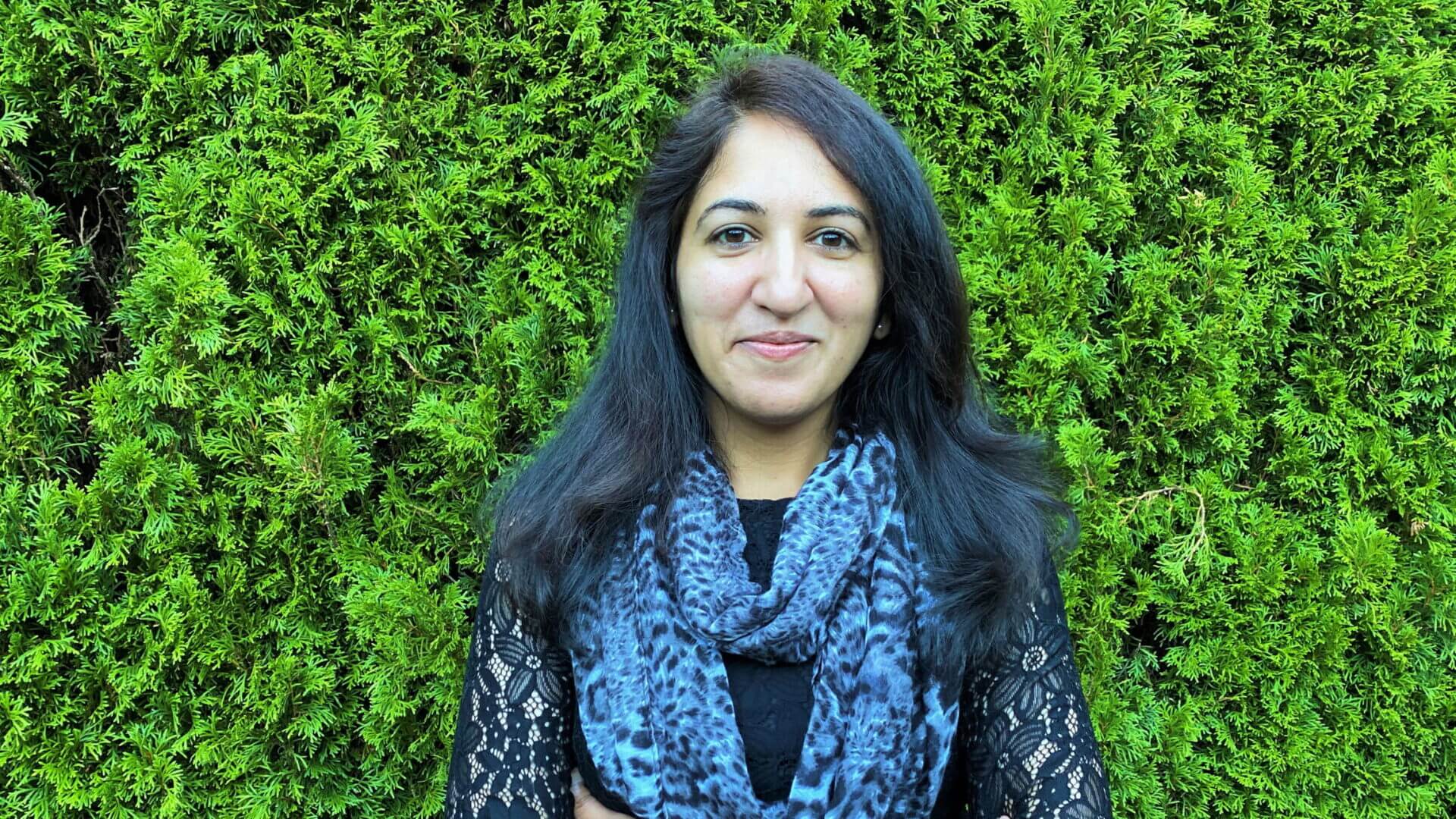 Gowri Krishnan stands in front of a green shrub with her arms crossed and smiles.