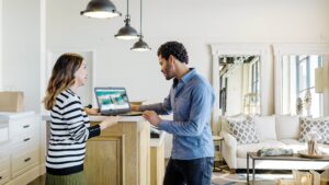 Female employee in commercial retail store providing customer support for male shopper. They are standing at a PoS counter in home furnishings showroom, looking at an HP laptop on the counter.