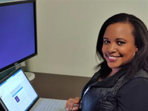 A smiling Chepkwony sits at a desk in a home office working on a laptop.