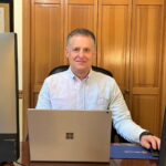 Pete Fortman is sitting at a desk in his home office in a blue-striped collared shirt. A laptop and two monitors with no information displayed are visible.