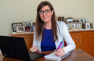 Tait, wearing glasses and a white sweater, sits at a wood desk with her laptop open and a notepad at the ready.