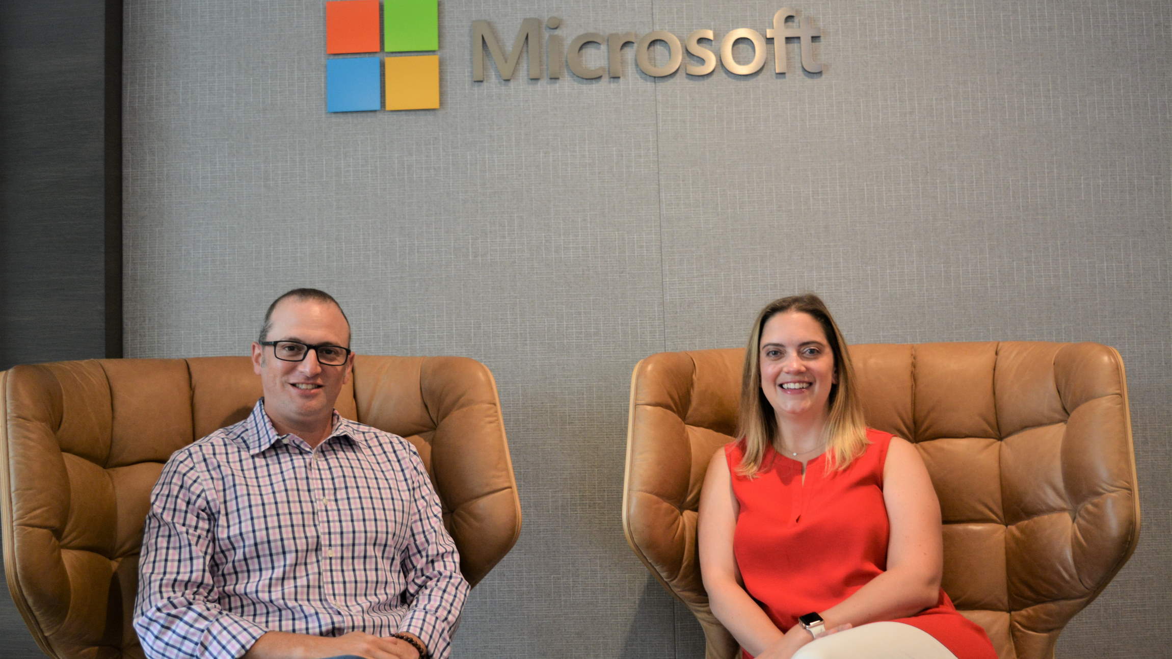 Yedinak and Laves sit and smile below a Microsoft logo inside a Microsoft building.