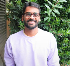 Rohun Patel, wearing glasses and a sweatshirt, stands outside in front of a large shrub.