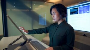 A woman uses a large, touchscreen computer in an open office setting.