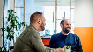 Side profile of male employee sitting next to coworker at table in cafeteria area of office.