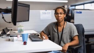 Portrait of female developer in glasses and short sleeved gray V neck t-shirt, facing camera.