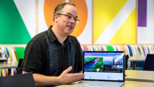 Sam Crewdson sits at a table in a cafe demoing the SharePoint Online modern experience on his Surface Book.