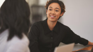 A woman smiles as she talks to another Microsoft employee while sitting a desk in an open working space.