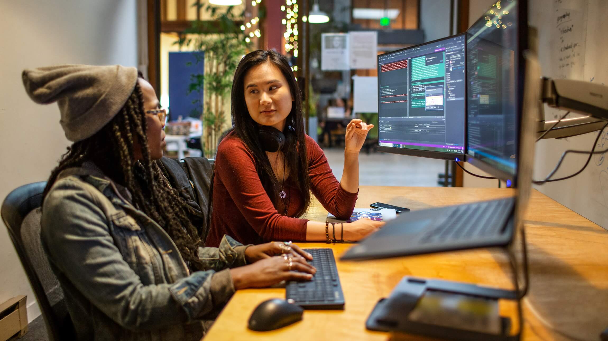 Two women talk to each other in front of computer screens with code displayed.