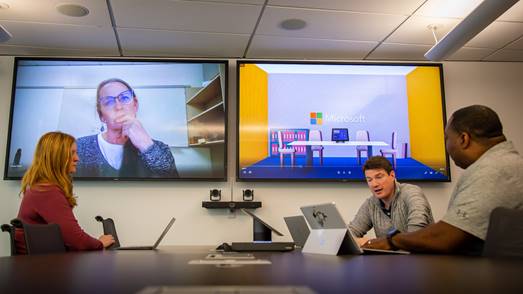 Photo of a dual-screen system in a conference room. On one screen is a live image of a meeting participant. The other screen shows content related to the meeting.