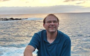 Johnson is outside facing the camera smiling, the sun is setting over the ocean in the background.