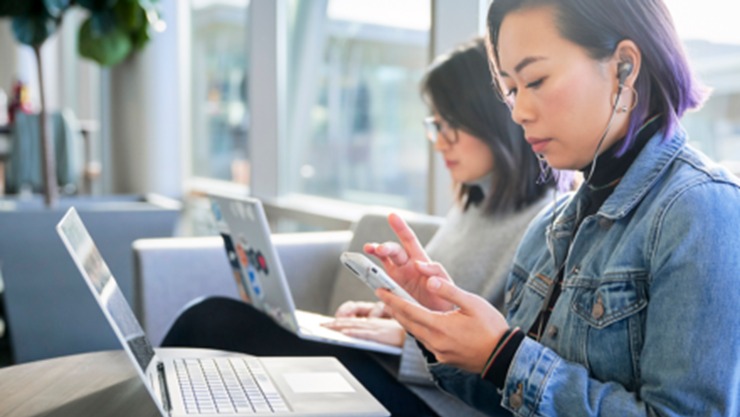 Two women sharing a couch. Each has a laptop in their lap. One woman is working her cell phone.