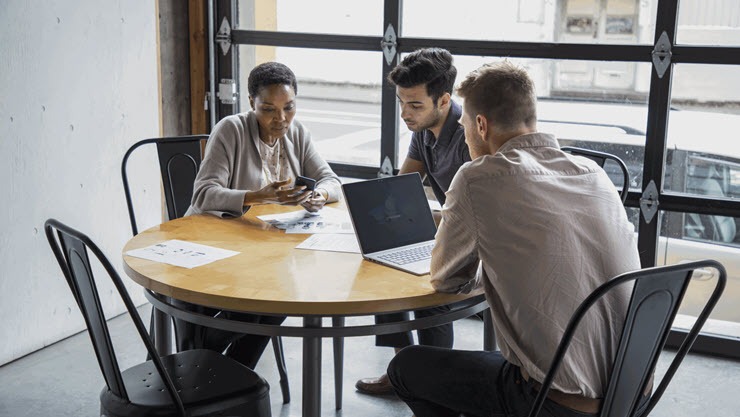 One female and two male workers sitting at round table in casual office setting or café/coffeehouse. The woman is holding a phone to show something to one of the men. The other man is using a Surface Laptop. 