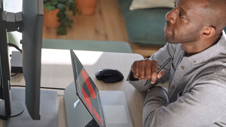 Adult male sitting at a desk holding a Surface Slim Pen 2 while looking at a large monitor.