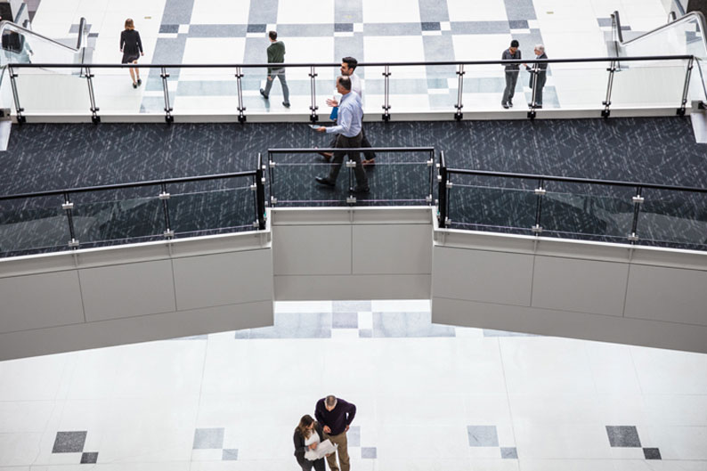 Aerial view of office building‘s interior entrance hall. Close-up of two separate groups of people standing together on an elevated walkway, chatting. Two others are walking on ground floor below.