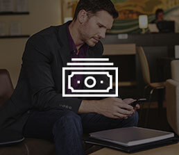 Bank customer in a waiting area checking his account on his phone with a money icon overlaid on the image.