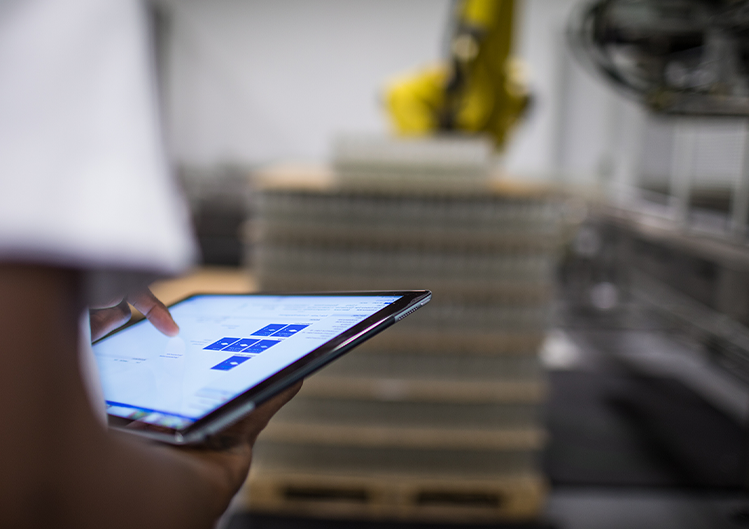 Close up of a woman holding a tablet in a manufacturing setting, with a machine robotic arm in the background.