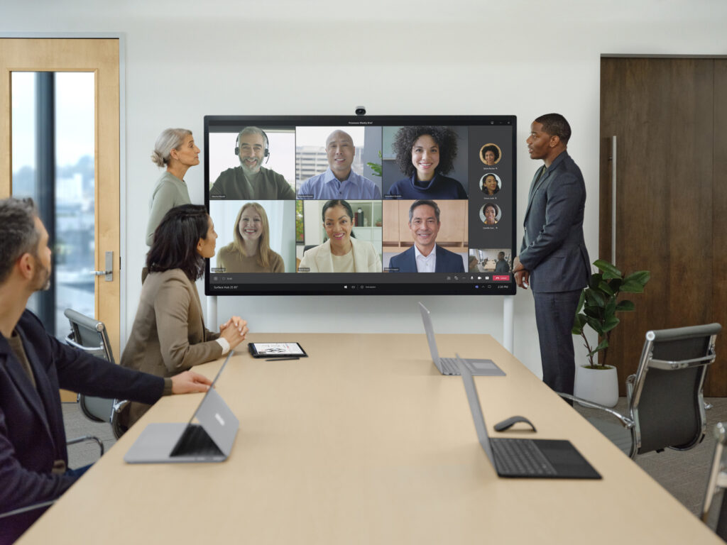 Dois colegas presentes em um Surface Hub 2 S durante uma teleconferência do Teams com membros de equipe presenciais e remotos.