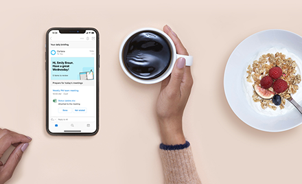 Cortana Daily Briefings on a mobile device. User holds a cup of coffee. Next to the coffee is a breakfast dish.